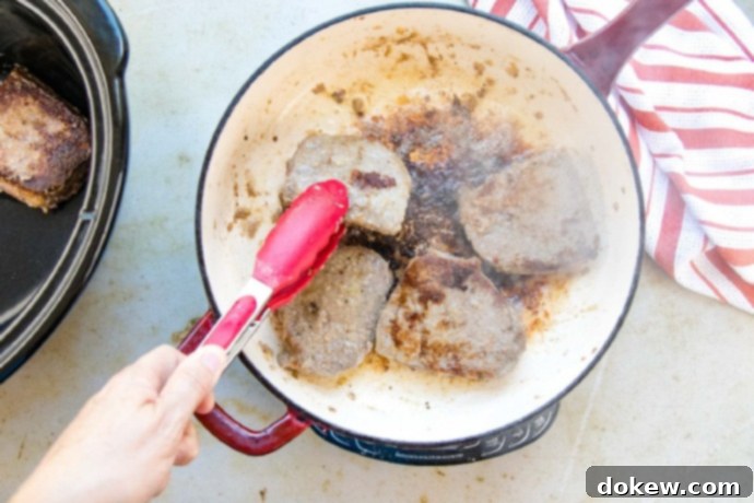 Savory Swiss Steak Cooked beef steaks being transferred from a skillet to a slow cooker.