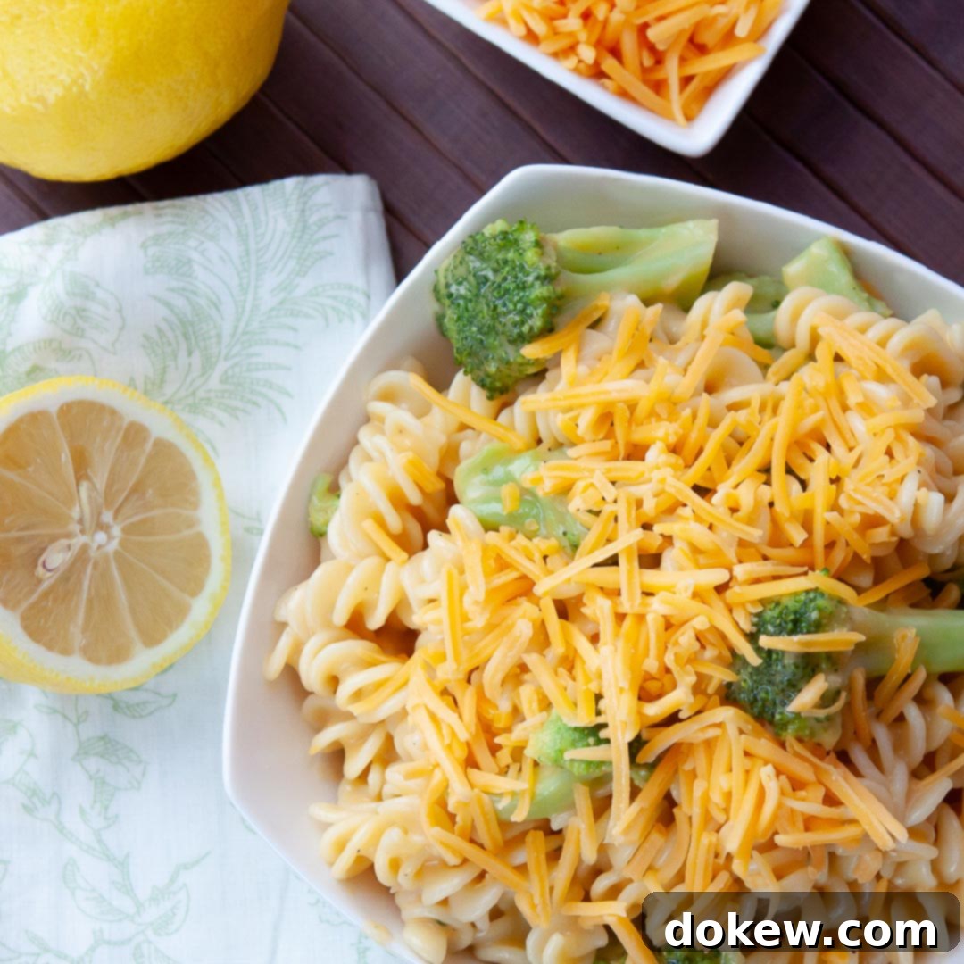 Cheddary Broccoli Spirals 2 White bowl with rotini pasta, broccoli, and shredded cheddar cheese, next to lemons and a bowl of shredded cheese
