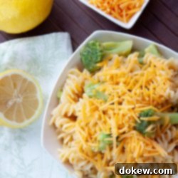Cheddary Broccoli Spirals 3 White bowl with rotini pasta, broccoli, and shredded cheddar cheese, next to lemons and a bowl of shredded cheese