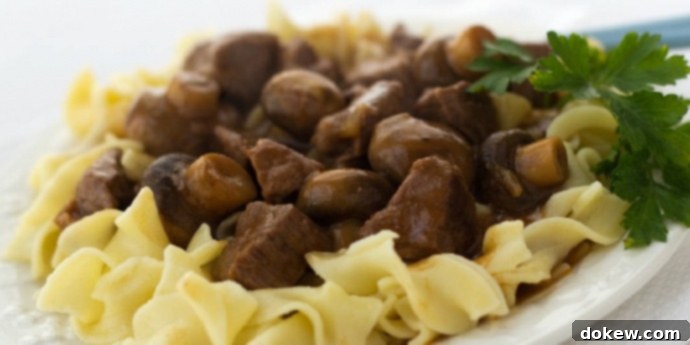 Slow Cooker Beef with Mushrooms A white plate generously topped with tender slow cooker beef and mushrooms, served over a bed of golden egg noodles and garnished with a sprig of fresh parsley, showcasing a comforting and hearty meal.