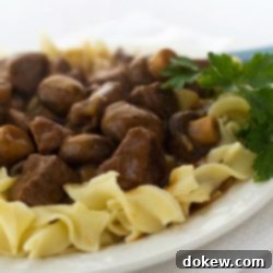 Hearty Slow Cooker Beef and Mushroom Stew 2 A smaller, cropped view of a white plate with egg noodles topped with slow cooker beef and mushrooms, garnished with a sprig of parsley.
