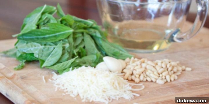 How to Make Homemade Pesto Photo is a pile of basil leaves, a glass measuring cup with olive oil, a pile of shredded parmesan cheese, garlic cloves, and a pile of pine nuts, on a wooden cutting board.