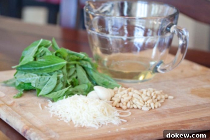 Ingredients for pesto: fresh basil, garlic, pine nuts, parmesan cheese, and olive oil a cutting board with a pile of basil leaves, cloves of garlic, a pile of pine nuts, a pile of shredded Parmesan cheese, and a measuring cup with oil