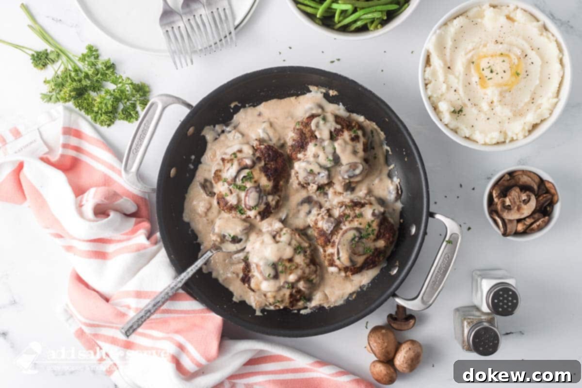 Several Salisbury Steak patties in mushroom cream sauce in a black pan with a serving spoon surrounded by salt and pepper shakers and bowls of green beans and mashed potatoes.