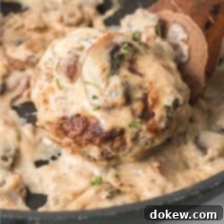 Rich Salisbury Steak in a Velvety Mushroom Sauce 7 Salisbury Steak patty in mushroom cream sauce in a black pan with a serving spoon.