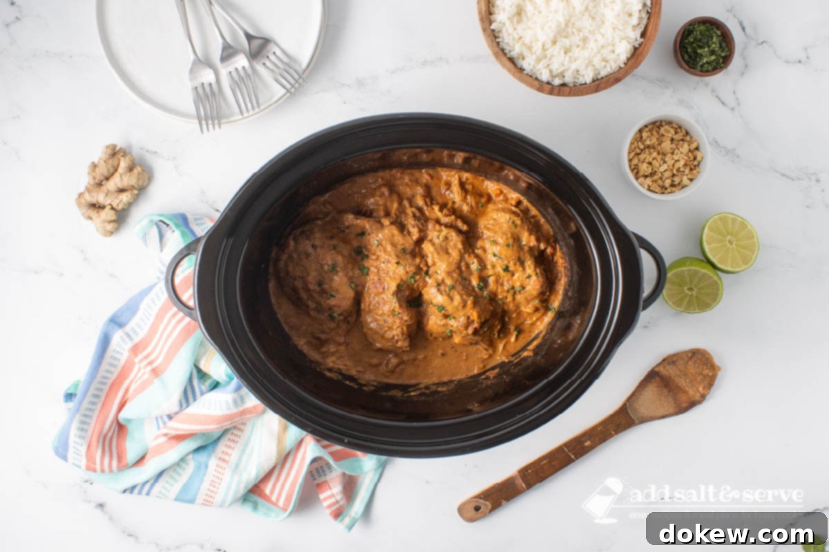 Weeknight Thai Peanut Chicken Magic 7 Cooked Thai Peanut Chicken in a slow cooker surrounded by rice, chopped peanuts, cilantro, ginger, a halved lime, and a stack of empty plates with forks.