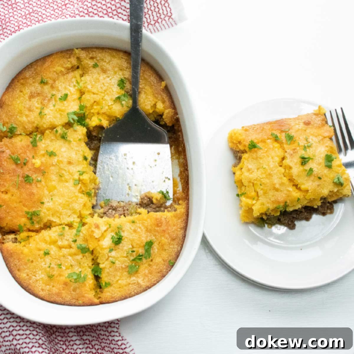 A delicious slice of Cornbread Burger Bake casserole on a white plate, with the full casserole dish visible in the background. A spatula rests in the dish, and a fork is on the plate, ready to enjoy this hearty meal.