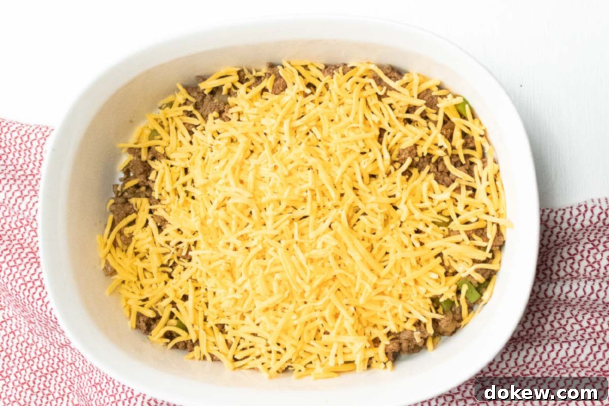 Cornbread Burger Casserole 4 A savory layer of cooked ground beef, diced onions, and bell peppers sits at the bottom of a white casserole dish, generously topped with an even layer of shredded cheddar cheese, ready for the cornbread topping.