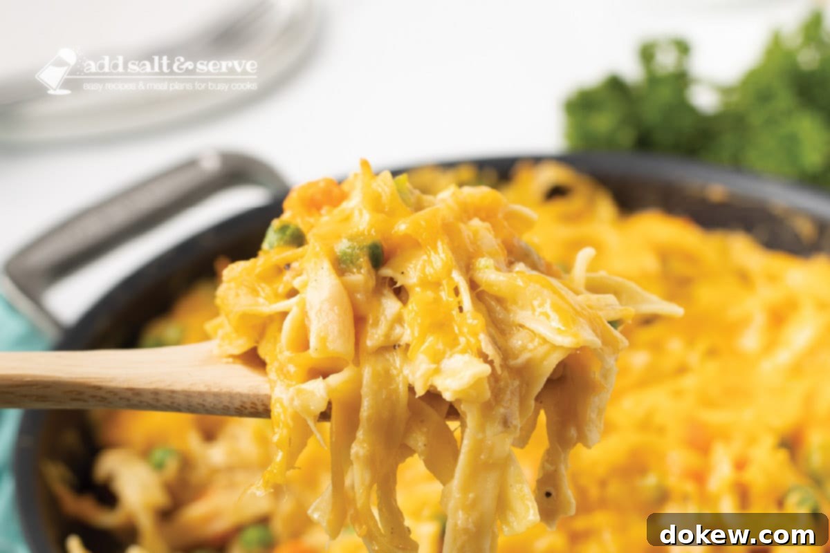 Comforting Tuna Noodle Bake 7 A spoonful of creamy and cheesy tuna casserole lifted from a skillet, showing the noodles, peas, and carrots.
