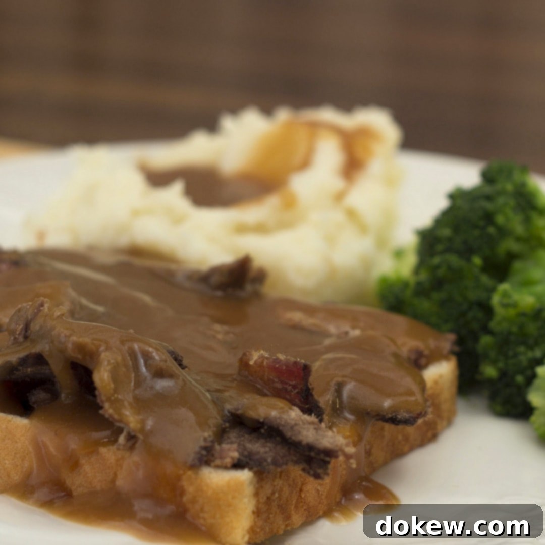 Photo of a slice of white bread with sliced roast beef and gravy, with mashed potatoes and gravy and cooked brocoli florets on a white plate