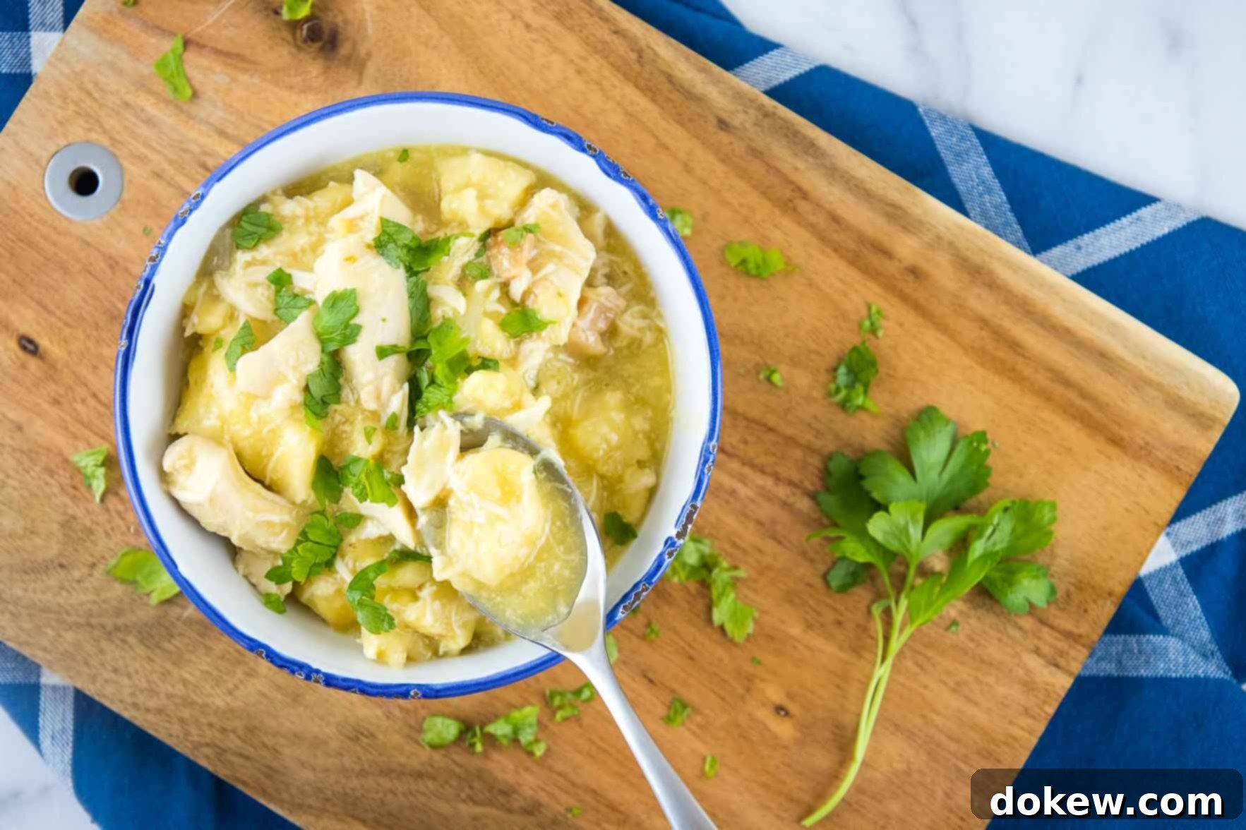 Hearty Slow Cooker Chicken and Biscuit Dumplings 3 Chicken and dumplings garnished with parsley in a white bowl with a blue rim. There is a spoon with a dumpling and some broth. The bowl is on a wooden cutting board on top of a blue napkin with white stripes, all on a white marble counter.