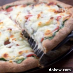 Aegean Feta & Olive Pizza 2 Serving spatula lifting a slice of pizza off of whole pizza on a pizza pan, pizza has thick crust, baby spinach onions, tomatoes, and mozzarella cheese.
