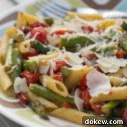 Fresh Tomato and Asparagus Carbonara 3 Cooked penne pasta, 1-inch pieces of asparagus, grape tomatoes, topped with a garnish of parmesan cheese on a white plate with blue and brown stripes