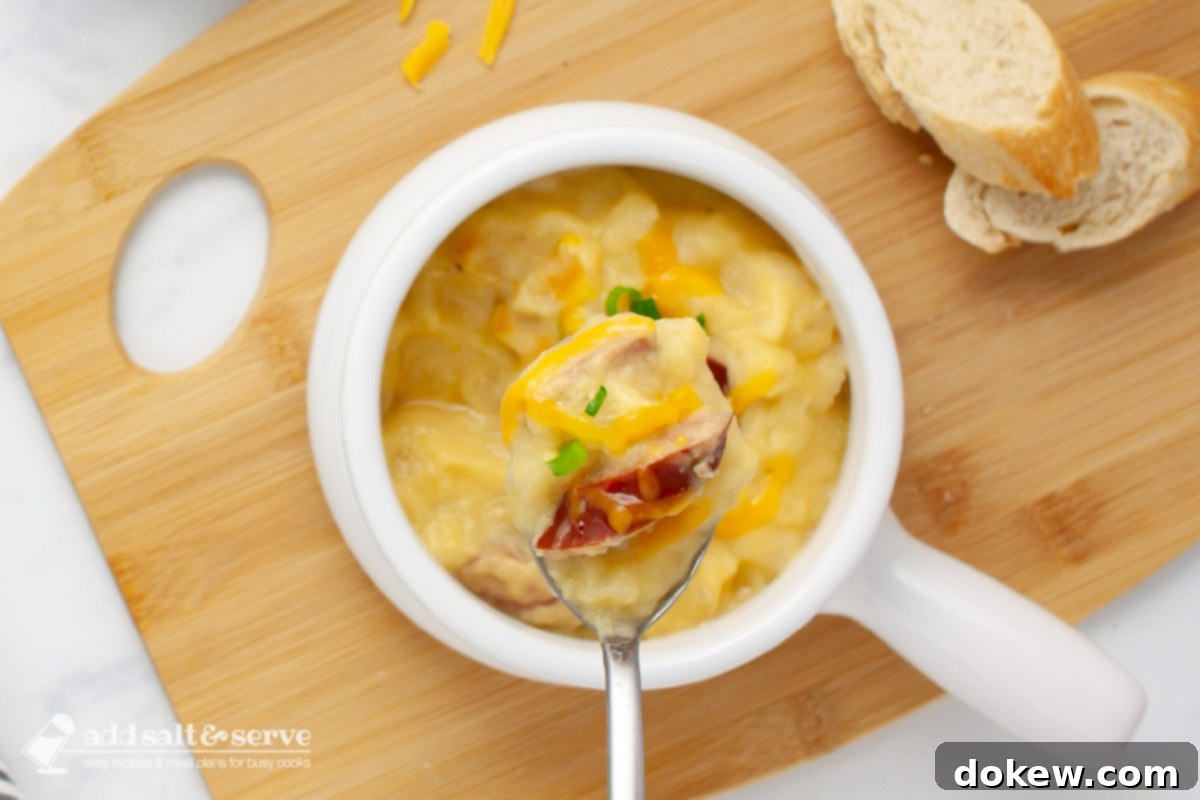 Hearty Cheesy Kielbasa Potato Slow Cooker Soup 2 Close-up of a spoonful of cheesy potato soup with kielbasa pieces garnished with shredded cheddar and sliced green onion on a spoon held over a crock of the soup, highlighting the creamy texture and delicious ingredients.
