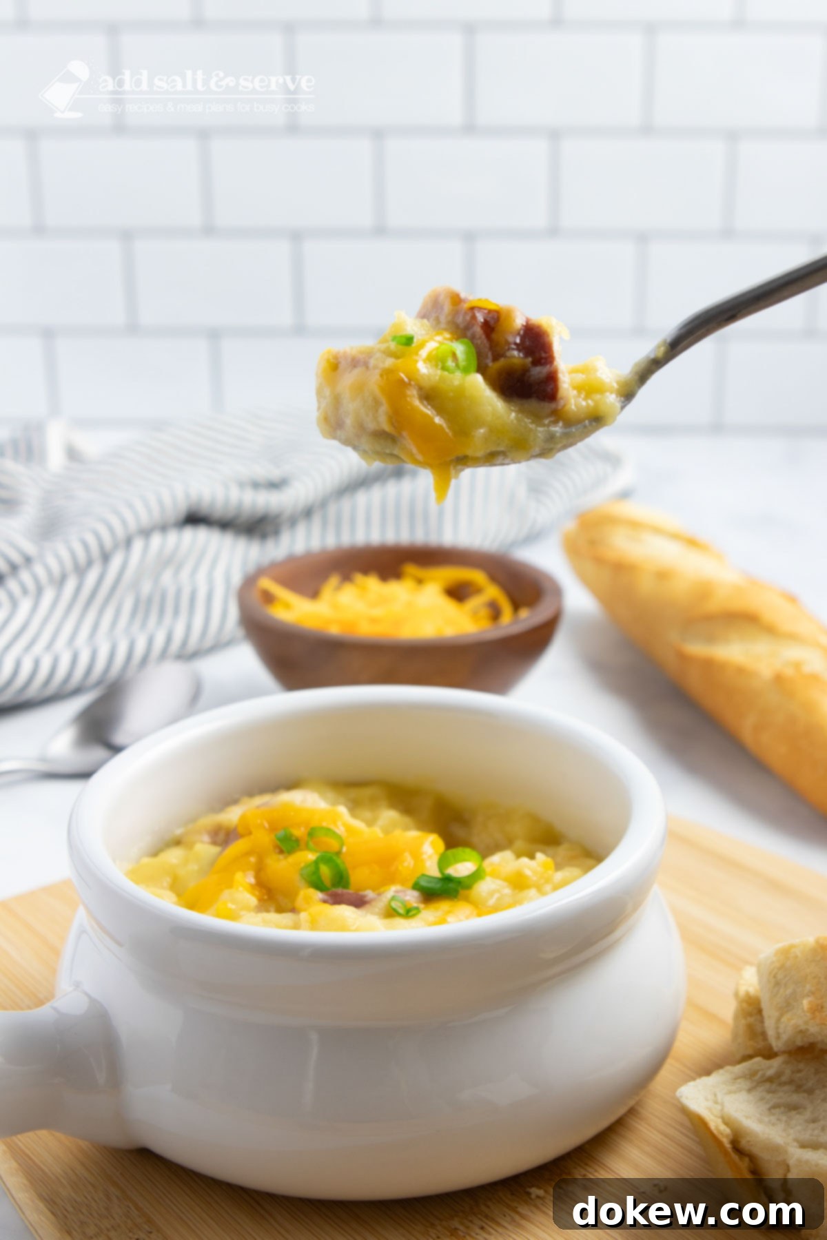 Hearty Cheesy Kielbasa Potato Slow Cooker Soup 6 Crock of cheesy potato soup garnished with shredded cheddar cheese and sliced green onion with a spoonful held above the crock with a baguette in the background, ready to be served.