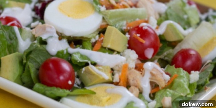 Cobb Salad with Chicken and Avocado: Fresh Ingredients A beautifully arranged Cobb salad on a white square plate, set against a cheerful yellow tablecloth. The salad features a bed of crisp lettuce, halved bright red grape tomatoes, perfectly sliced hard-boiled eggs, generous cubes of creamy avocado, tender diced chicken, vibrant shredded carrots, and a drizzle of rich blue cheese dressing.