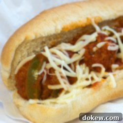 Authentic Homemade Italian Meatball Subs 2 Meatballs, sauce, shredded mozzarella cheese, and strips of bell peppers on a hot dog bun, all on a white plate.