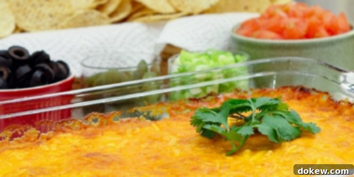 Nacho Bar: Customizable Family Meal Clear glass casserole dish with beans topped with melted cheese and garnished with a sprig of cilantro. In the background is a red bowl of sliced black olives, a clear bowl of jalapeño slices, a clear bowl of diced green onions, a white bowl of diced tomatoes, and a basket of tortilla chips, all ready for a customizable nacho bar.