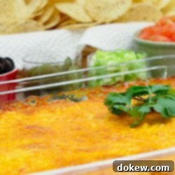 Nacho Station 2 Clear glass casserole dish with beans topped with melted cheese and garnished with a sprig of cilantro. In the background is a red bowl of sliced black olives, a clear bowl of jalapeño slices, a clear bowl of diced green onions, a white bowl of diced tomatoes, and a basket of tortilla chips.
