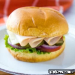 Turkey and Swiss Hoagies 2 A close-up shot of a turkey and Swiss hoagie on a brioche bun, revealing the rich layers of deli turkey, melted Swiss cheese, fresh lettuce, tomato, pickle, and onion. The sandwich is presented on a white and orange plate, with a stylish coral and white windowpane towel blurred in the background.