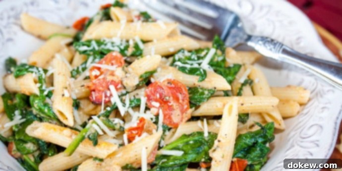Pasta Carbonara with Tomatoes and Spinach A beautifully presented white plate filled with perfectly cooked penne pasta, vibrant sautéed baby spinach, glistening halved cherry tomatoes, and a generous sprinkle of shredded mozzarella cheese, with a fork resting gracefully on the side.