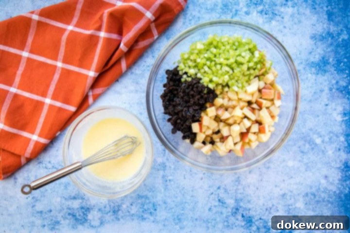 apple-raisin-salad-ip-2-min Overhead view of a large bowl with chopped apples, raisins, and celery, a smaller bowl with a creamy salad dressing and a whisk, all on a blue surface with a coral plaid towel to the side