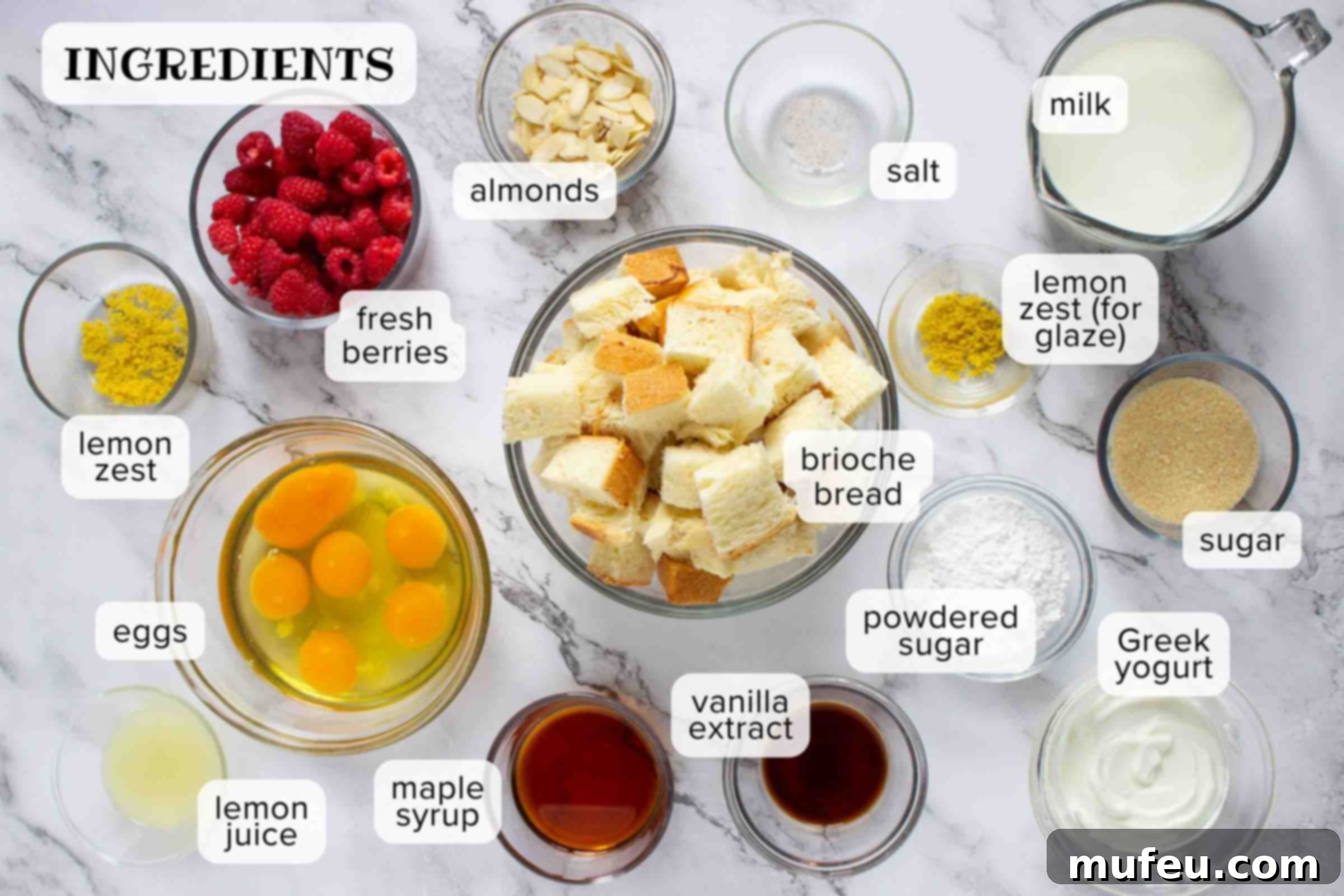 Ingredients on a counter: brioche bread, eggs, lemon juice, maple syrup, vanilla extract, powdered sugar, Greek yogurt, sugar, lemon zest, milk, salt, almonds, and berries.